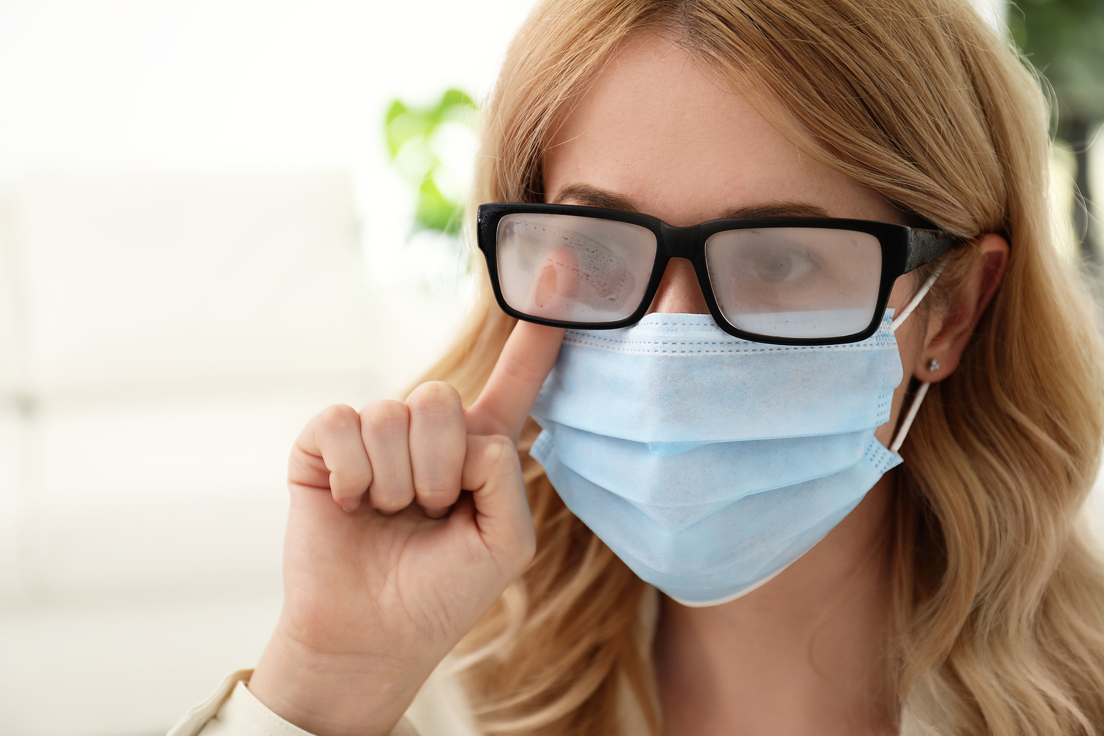 Woman wiping foggy glasses caused by wearing medical mask indoors, closeup